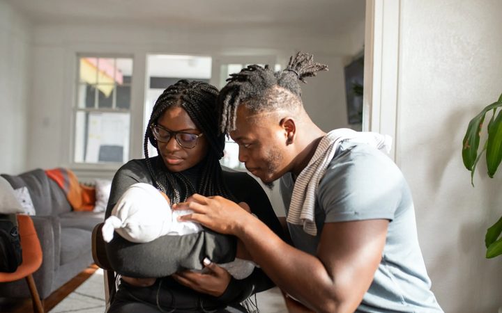 Two black parents cradling their swaddled newborn in a trendy apartment.
