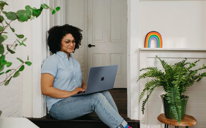 A person of color sitting in a doorway, with a few plants surrounding them, and a rainbow decoration on a nearby shelf.