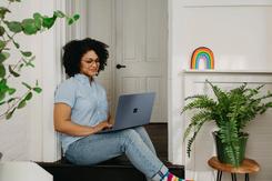 A person of color sitting in a doorway, with a few plants surrounding them, and a rainbow decoration on a nearby shelf.