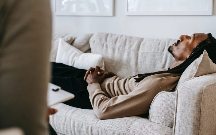 A black man with dreadlocks lies, despondent, on a gray loveseat before a therapist holding a clipboard.