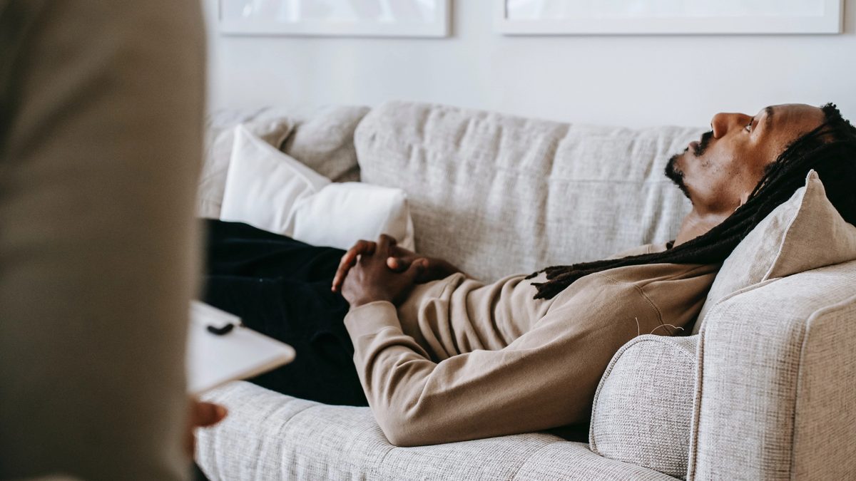 A black man with dreadlocks lies, despondent, on a gray loveseat before a therapist holding a clipboard.