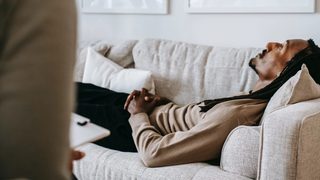 A black man with dreadlocks lies, despondent, on a gray loveseat before a therapist holding a clipboard.