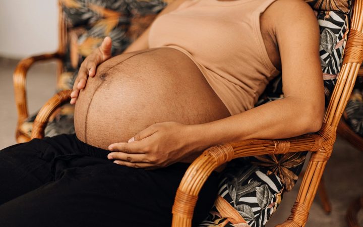 An exhausted, Black pregnant woman reclines in a chair made of cane, exposing their baby bump and cradling their stomach.