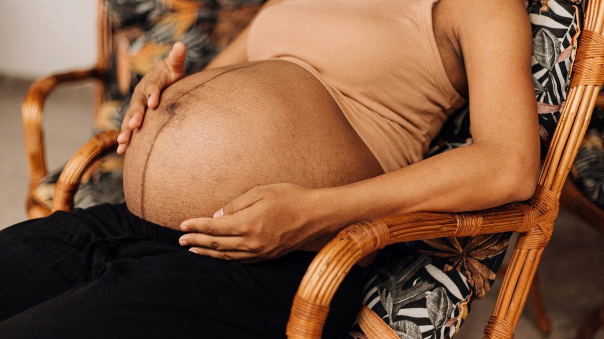 An exhausted, Black pregnant woman reclines in a chair made of cane, exposing their baby bump and cradling their stomach.