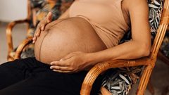 An exhausted, Black pregnant woman reclines in a chair made of cane, exposing their baby bump and cradling their stomach.