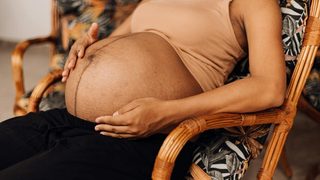 An exhausted, Black pregnant woman reclines in a chair made of cane, exposing their baby bump and cradling their stomach.