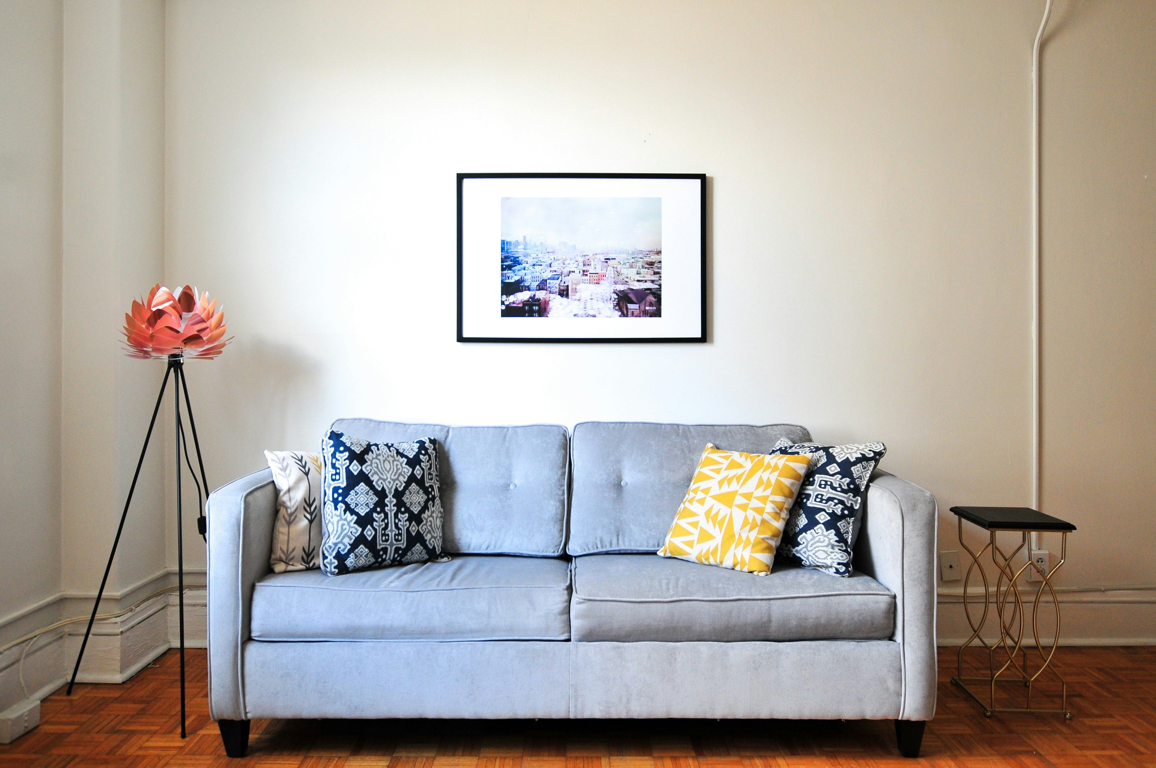 A light gray couch with ornate blue and yellow pillows, with a framed piece of art on the wall and a floral decoration on the left side.