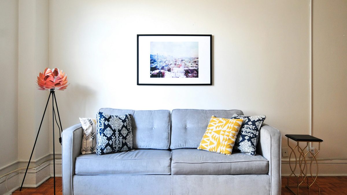 A light gray couch with ornate blue and yellow pillows, with a framed piece of art on the wall and a floral decoration on the left side.