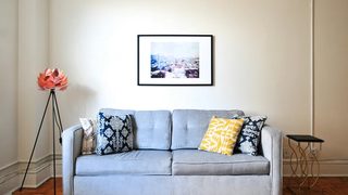 A light gray couch with ornate blue and yellow pillows, with a framed piece of art on the wall and a floral decoration on the left side.