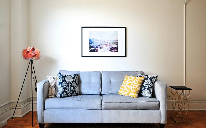 A light gray couch with ornate blue and yellow pillows, with a framed piece of art on the wall and a floral decoration on the left side.