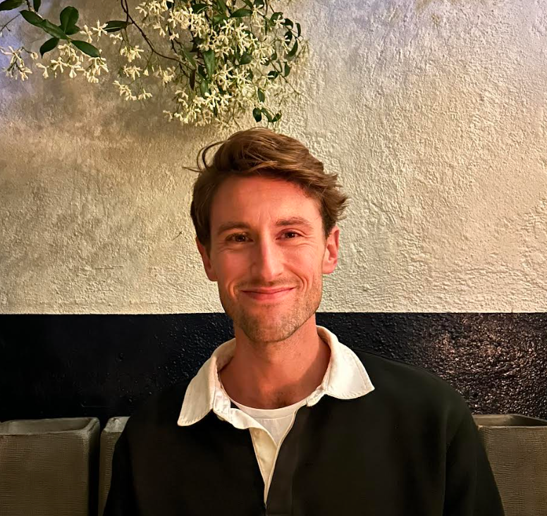 The author, Ryan, with short hair and a slight grin, sits against a striped backdrop with a floral plant hanging over his head.