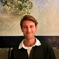 The author, Ryan, with short hair and a slight grin, sits against a striped backdrop with a floral plant hanging over his head.