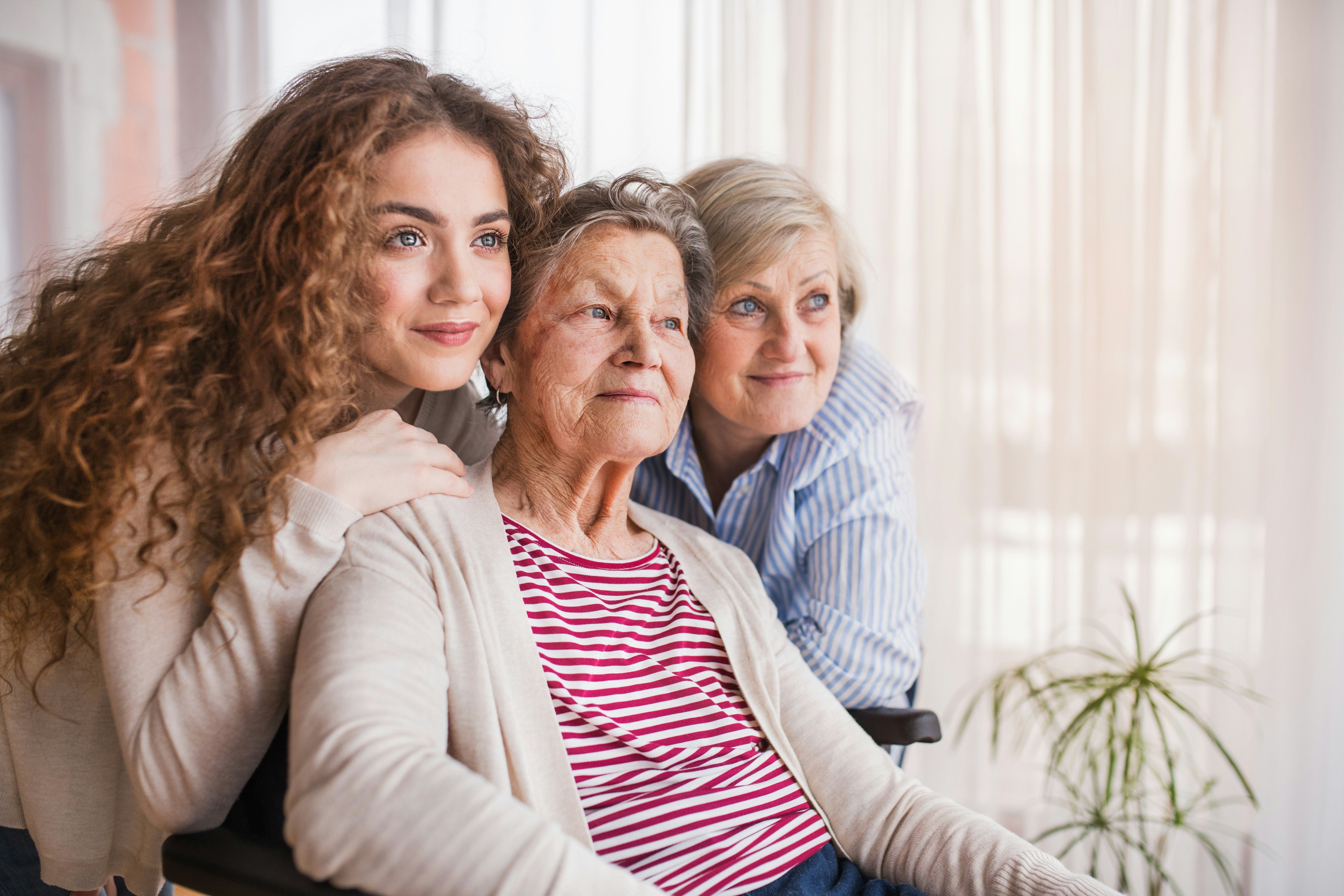 Three generations of women smiling together, with the oldest woman seated in a wheelchair.