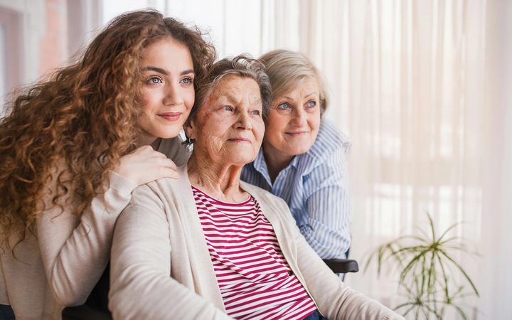 Three generations of women smiling together, with the oldest woman seated in a wheelchair.