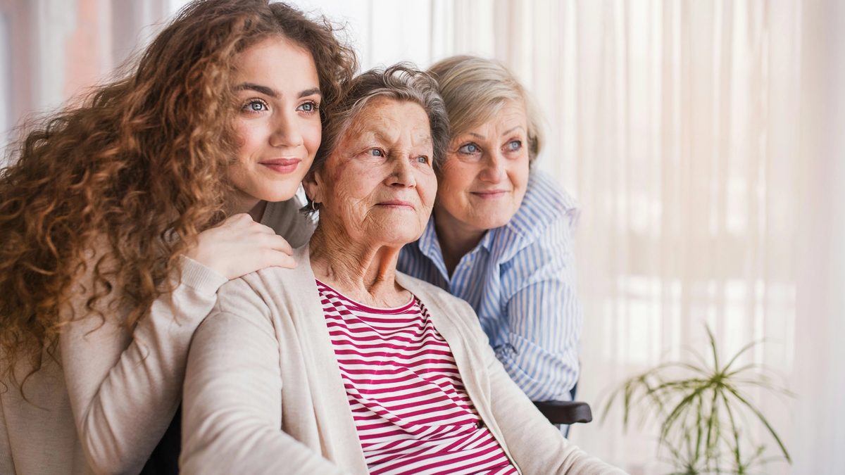 Three generations of women smiling together, with the oldest woman seated in a wheelchair.