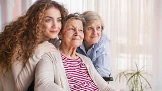 Three generations of women smiling together, with the oldest woman seated in a wheelchair.