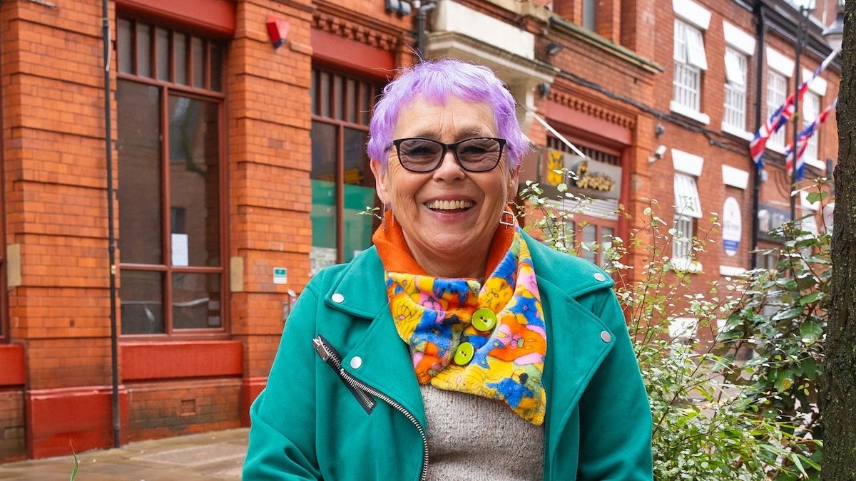 An older woman with purple hair and brightly colored clothes stands smiling confidently in front of a brick building in the city