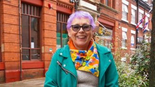 An older woman with purple hair and brightly colored clothes stands smiling confidently in front of a brick building in the city