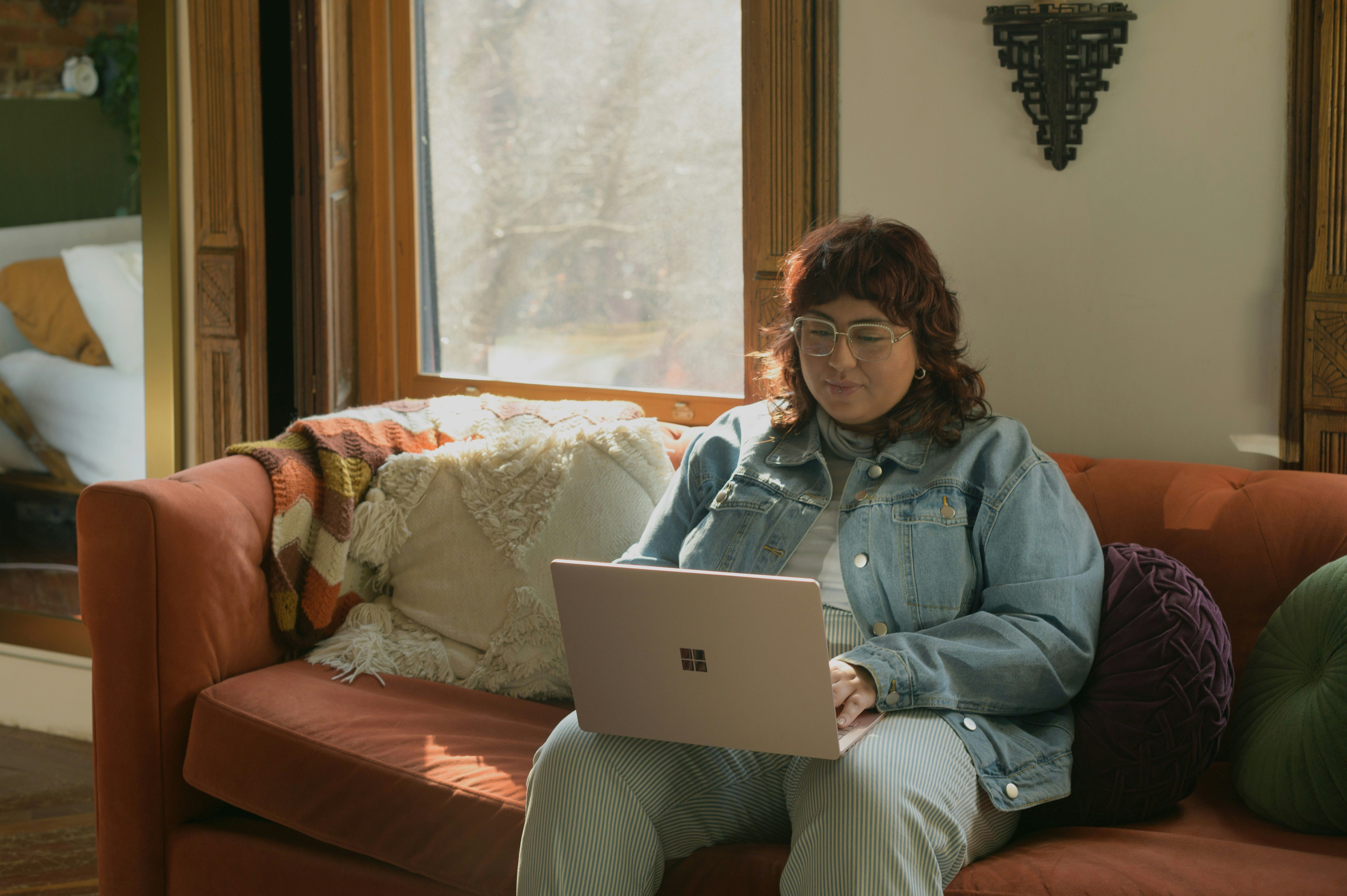 A person with long red hair sits on a red couch, dressed in all denim, looking down at a laptop.