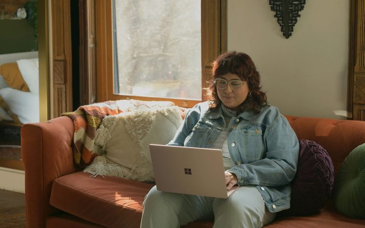 A person with long red hair sits on a red couch, dressed in all denim, looking down at a laptop.
