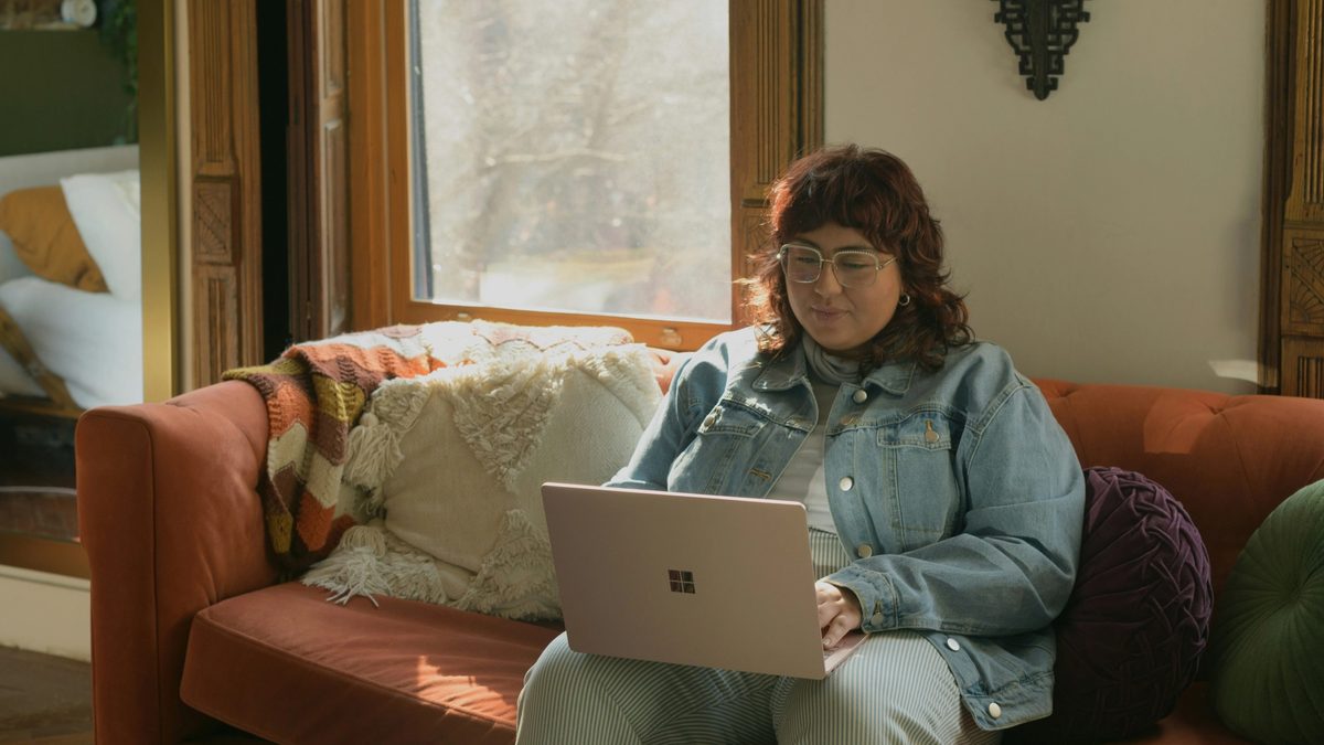 A person with long red hair sits on a red couch, dressed in all denim, looking down at a laptop.