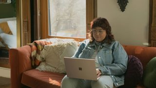 A person with long red hair sits on a red couch, dressed in all denim, looking down at a laptop.