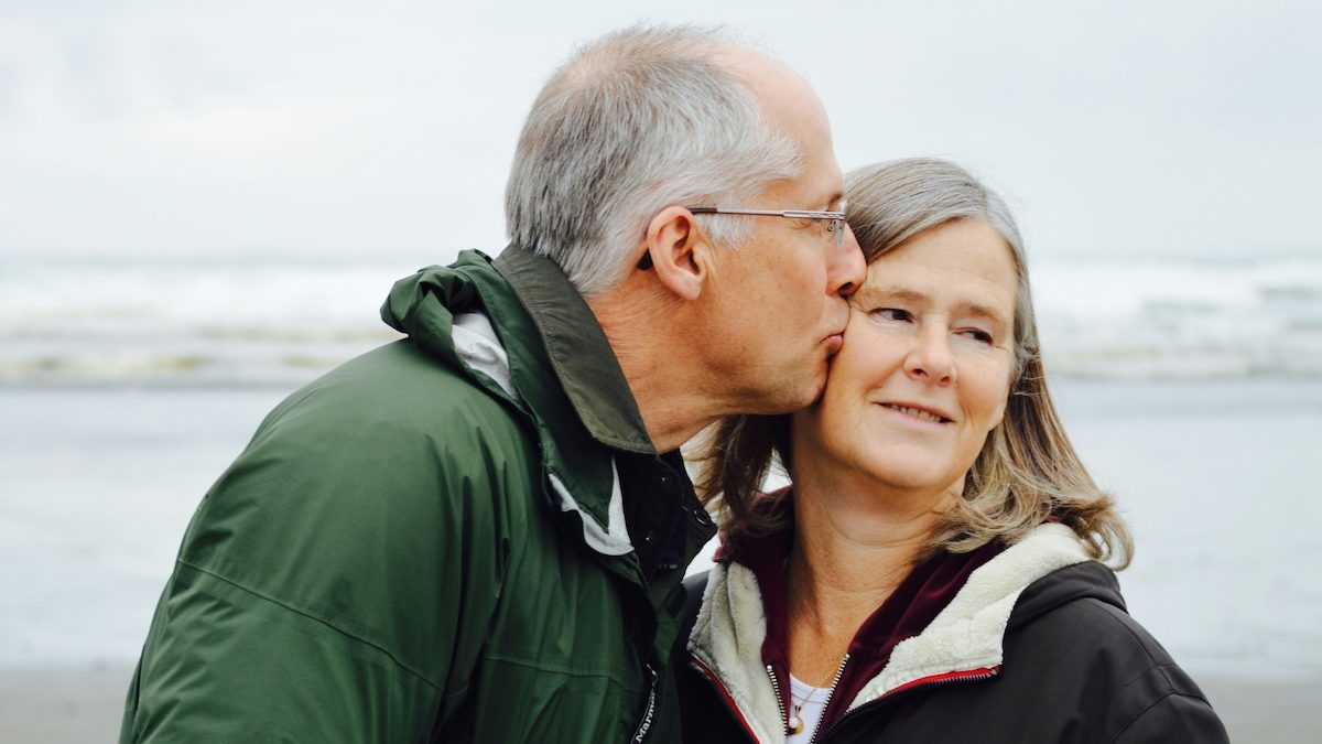 An older man in a green jacket kisses his wife's cheek on the beach