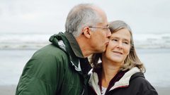 An older man in a green jacket kisses his wife's cheek on the beach
