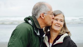 An older man in a green jacket kisses his wife's cheek on the beach