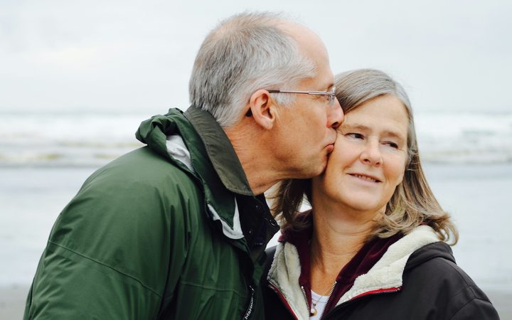 An older man in a green jacket kisses his wife's cheek on the beach