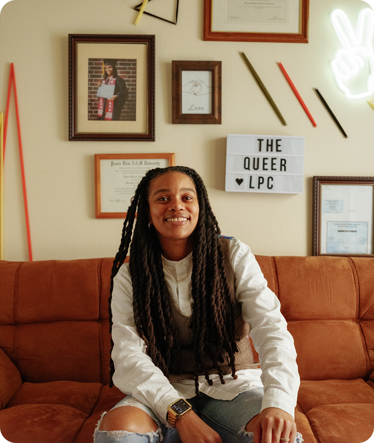 A provider sitting on a couch smiling in front of a wall of pictures, certificates, and a sign that says The Queer LPC