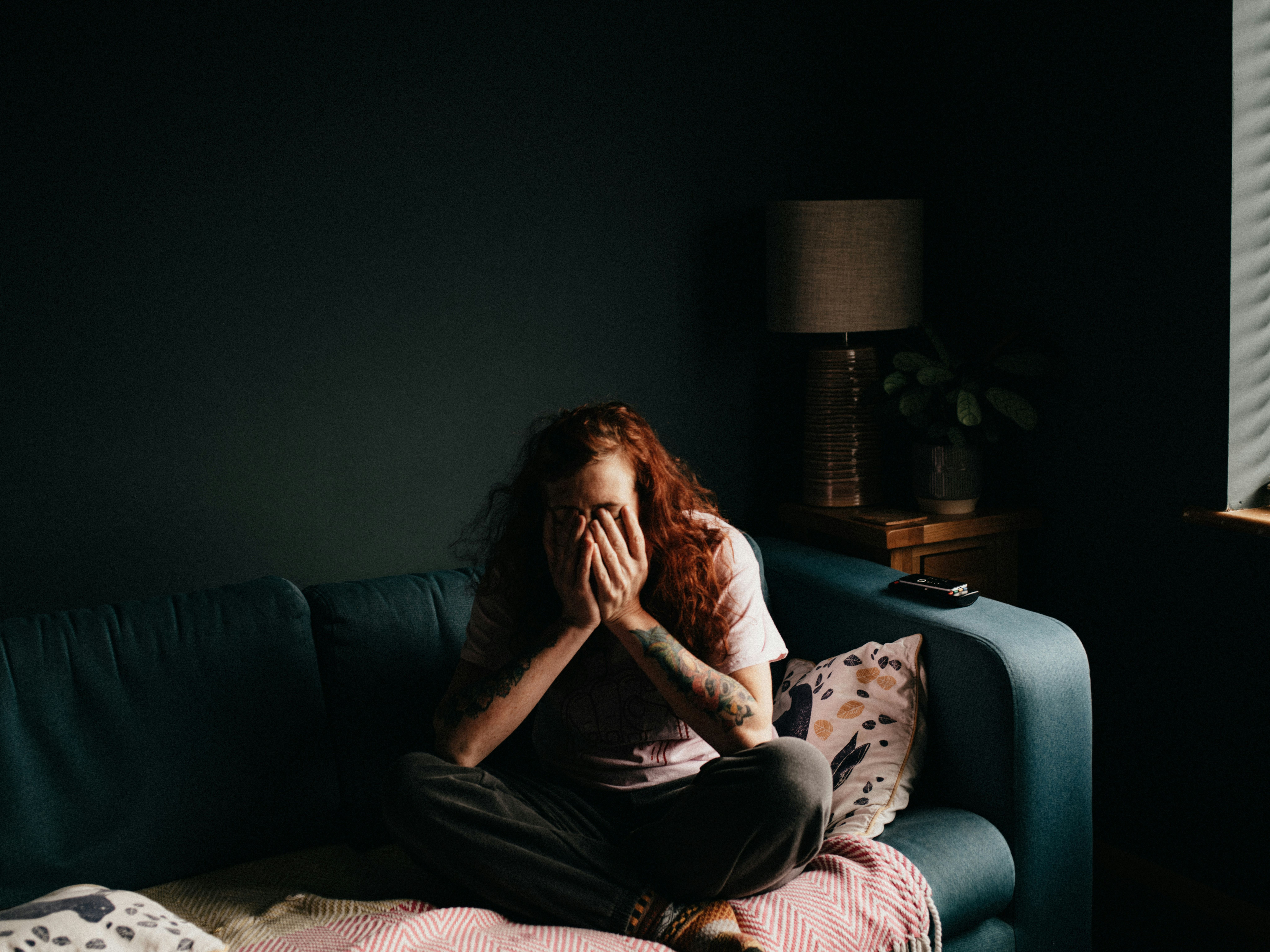 A person with long, red, curly hair sits cross-legged on a teal couch in a dimly-lit room, covering their face with their hands.