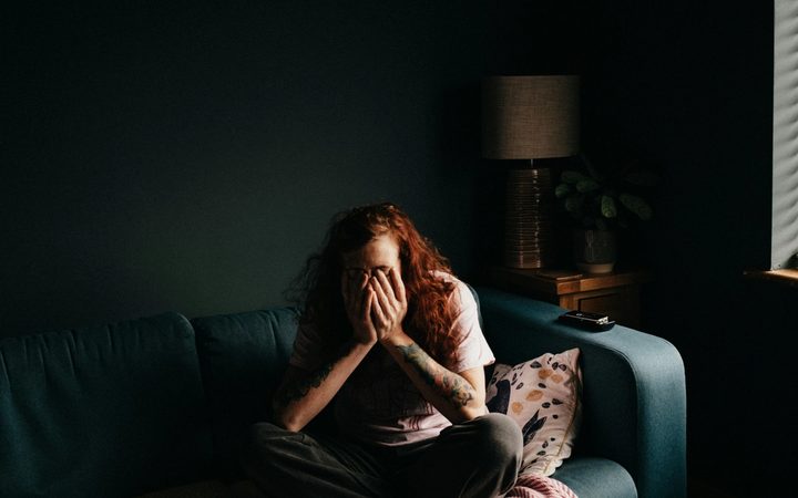 A person with long, red, curly hair sits cross-legged on a teal couch in a dimly-lit room, covering their face with their hands.