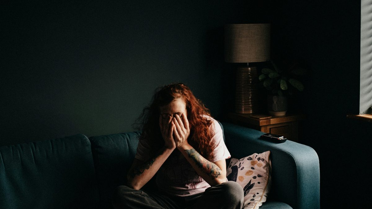 A person with long, red, curly hair sits cross-legged on a teal couch in a dimly-lit room, covering their face with their hands.
