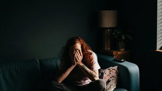 A person with long, red, curly hair sits cross-legged on a teal couch in a dimly-lit room, covering their face with their hands.