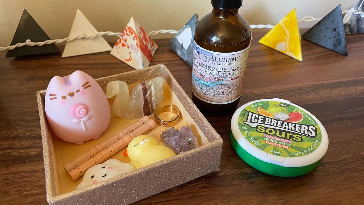 A small tray of fidget toys, including a pink cat, orange magnets, a purple crystal, and a rose gold spinning ring, as well as a spray bottle of essential oils and a green tin of sour candies. There are small paper lanterns lining the background.