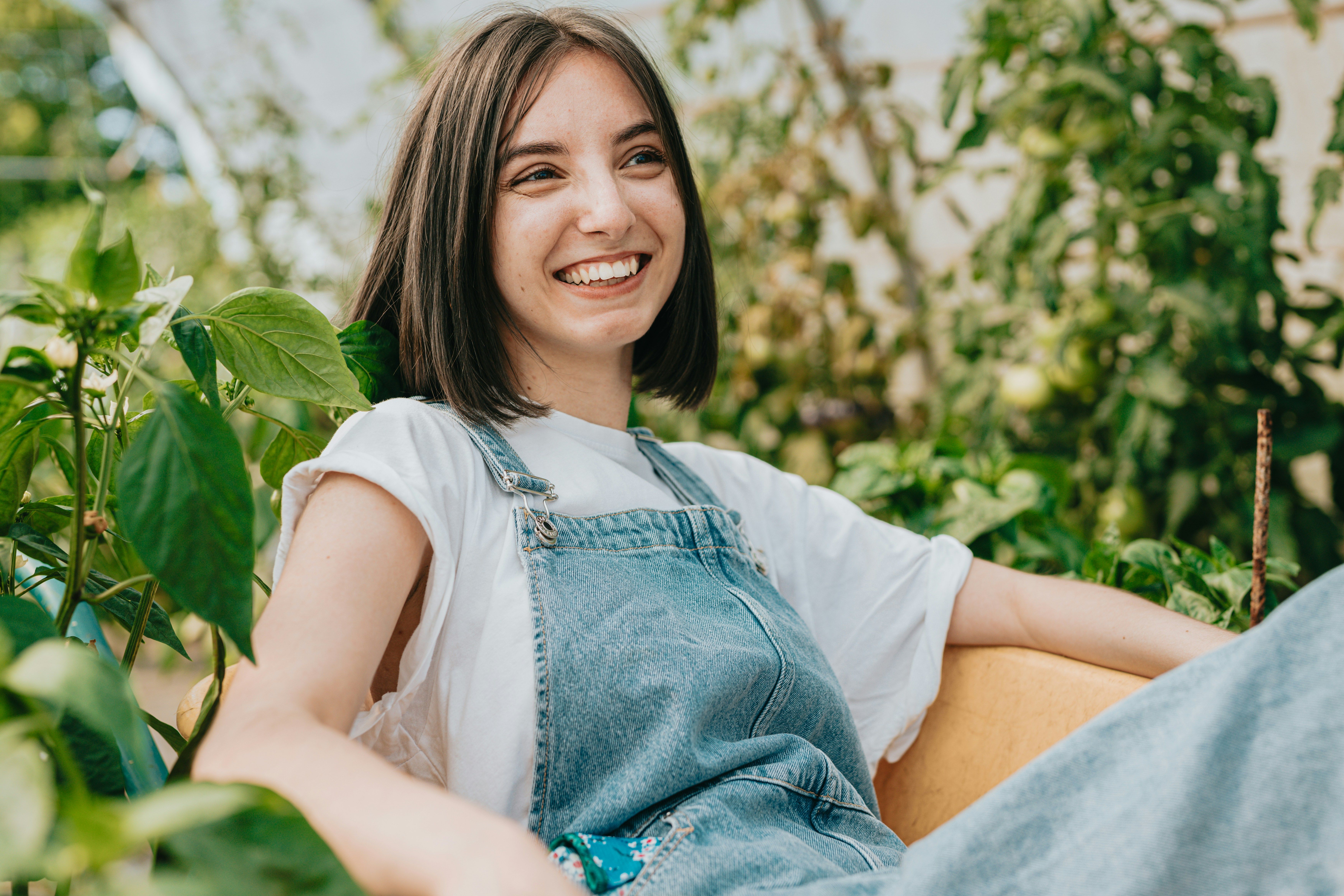 Smiling young woman in denim overalls sitting outdoors among green plants
