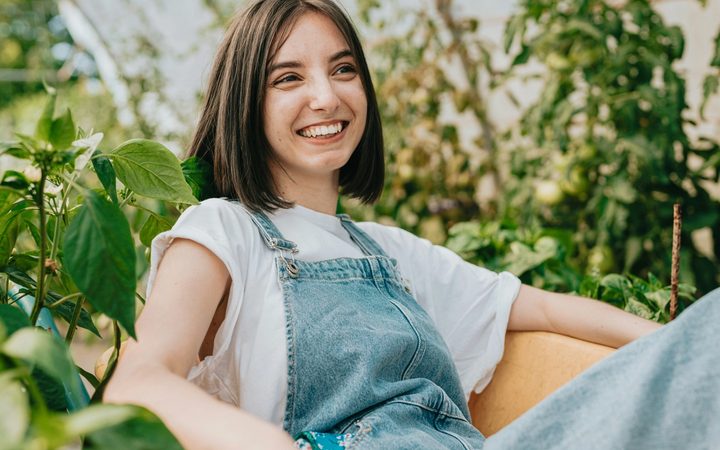 Smiling young woman in denim overalls sitting outdoors among green plants