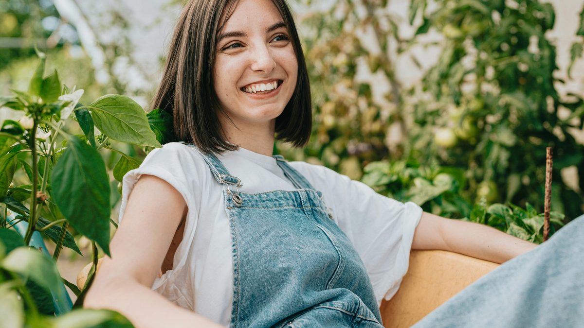 Smiling young woman in denim overalls sitting outdoors among green plants