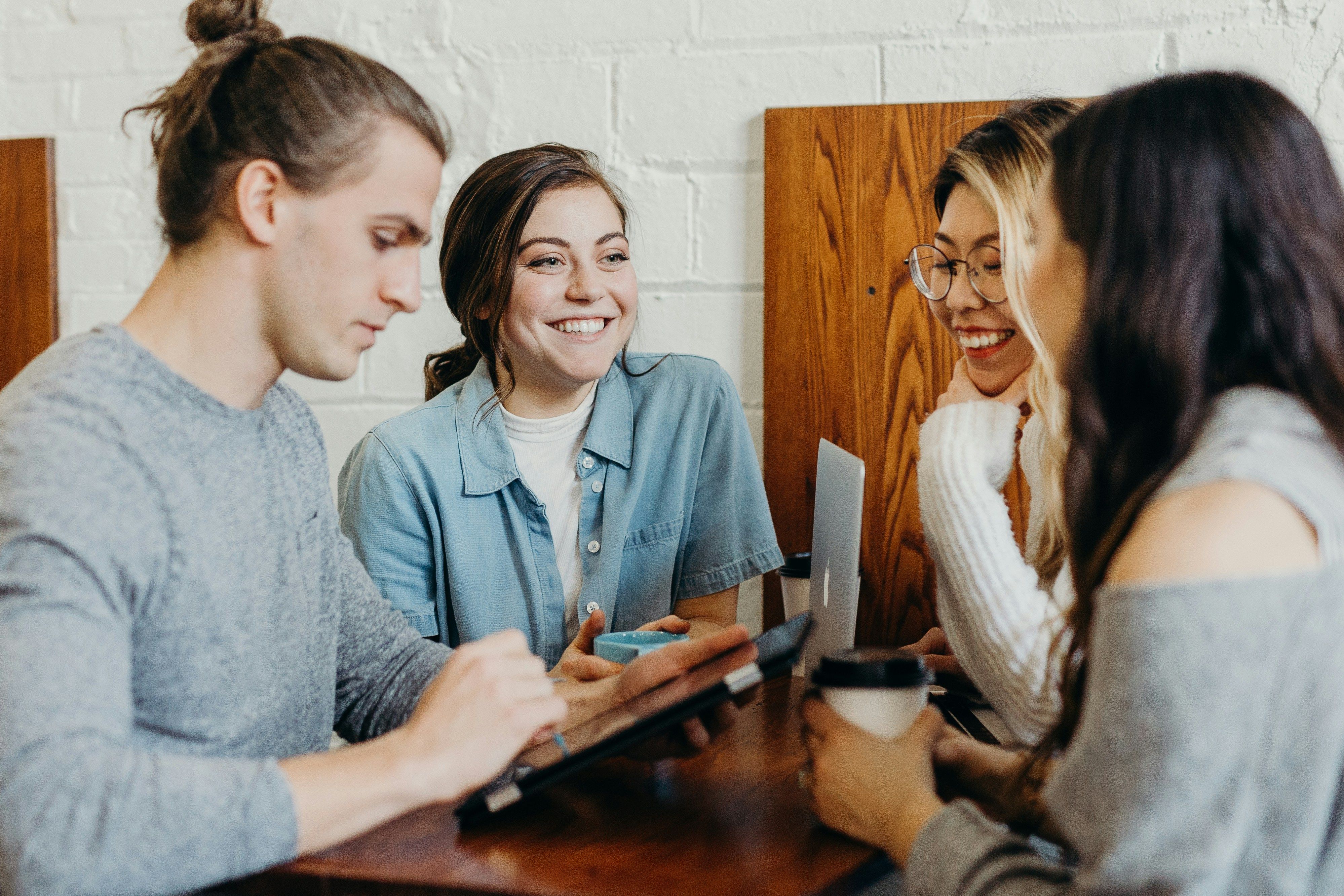 Four individuals sit around a table, engaged in conversation. One person is looking down at a tablet, another is looking up and smiling at someone across the table, another is smiling and looking down, and another is holding a to-go coffee cup.