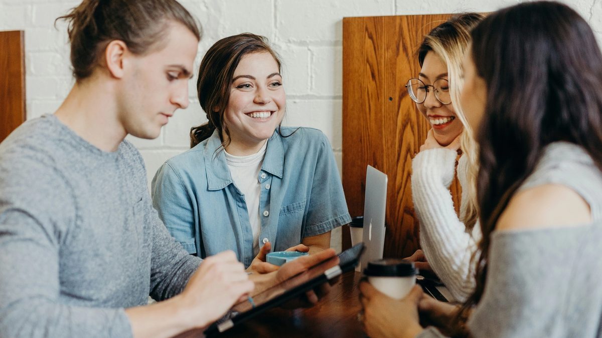 Four individuals sit around a table, engaged in conversation. One person is looking down at a tablet, another is looking up and smiling at someone across the table, another is smiling and looking down, and another is holding a to-go coffee cup.