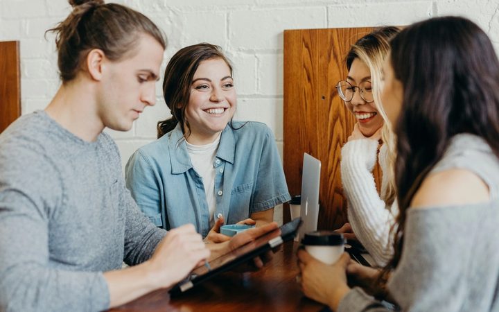 Four individuals sit around a table, engaged in conversation. One person is looking down at a tablet, another is looking up and smiling at someone across the table, another is smiling and looking down, and another is holding a to-go coffee cup.