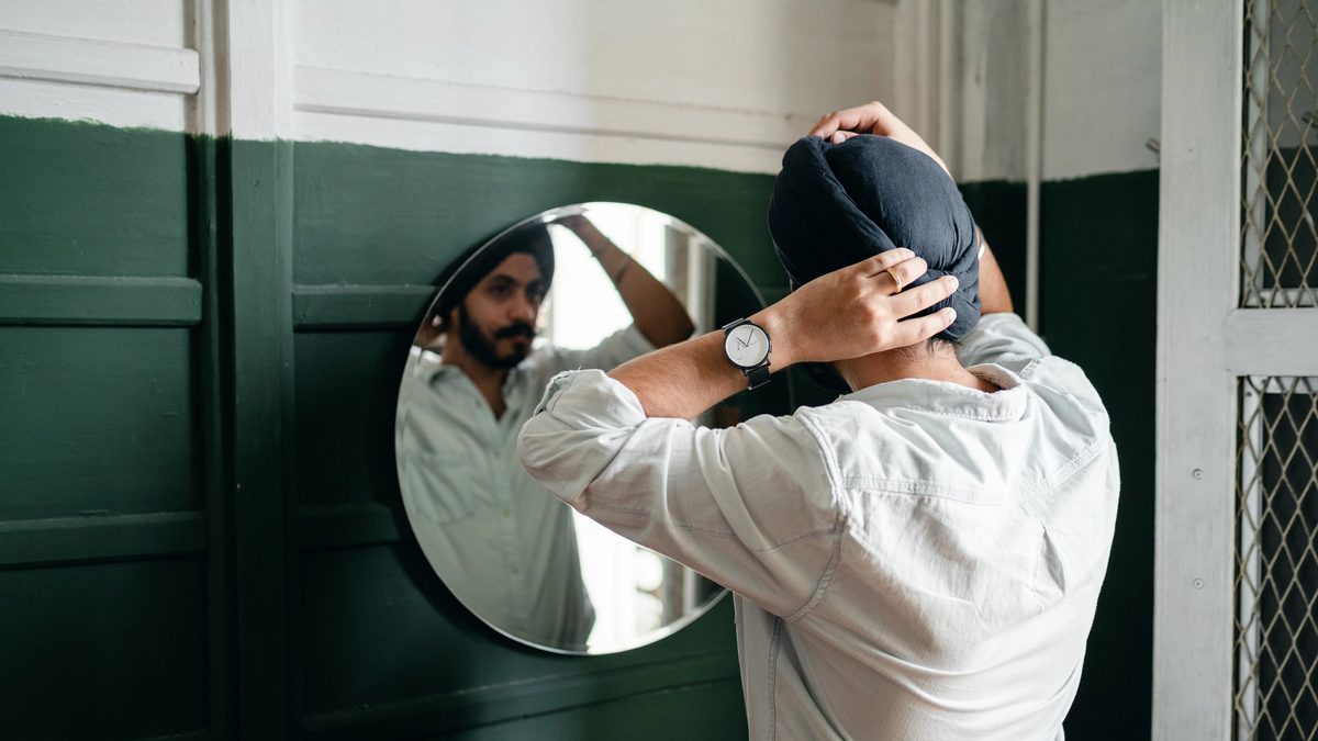 A Sikh man wearing a dastar straightens his turban in the mirror.