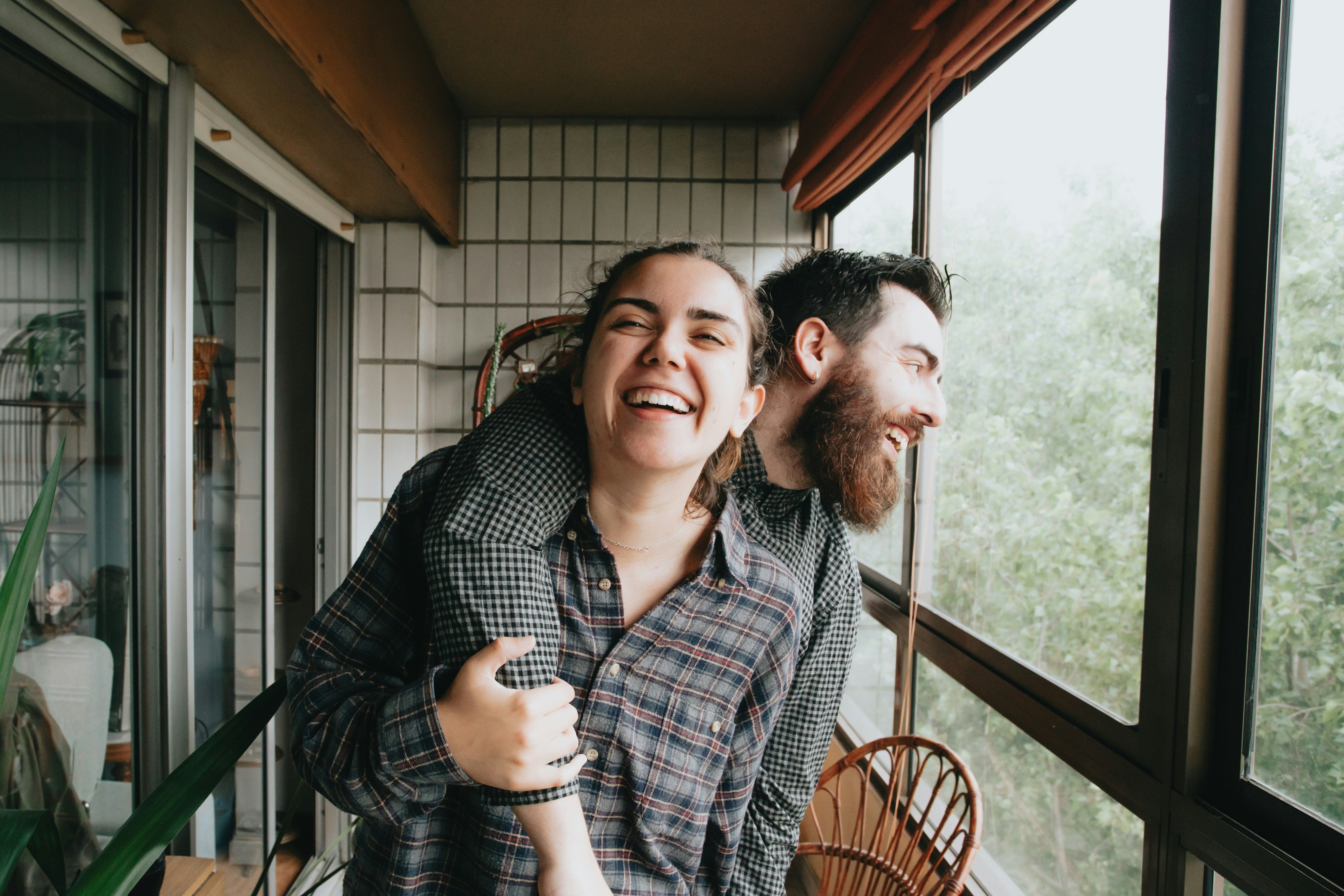 A happy couple stands in an enclosed sunroom overlooking some greenery. One stands laughing with their arm around the other, who is clutching the arm and smiling in return. Both appear to be laughing.