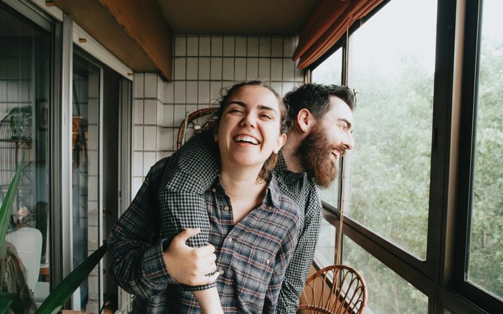 A happy couple stands in an enclosed sunroom overlooking some greenery. One stands laughing with their arm around the other, who is clutching the arm and smiling in return. Both appear to be laughing.