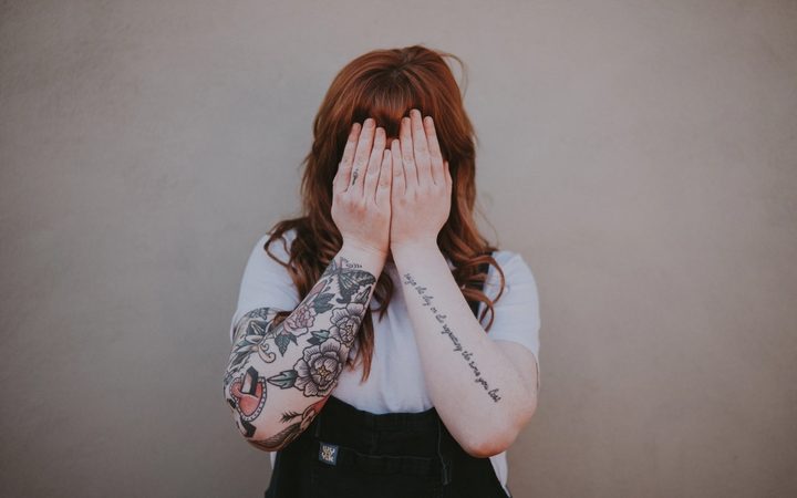 A person with long red hair and tattooed arms covers their face with their hands.