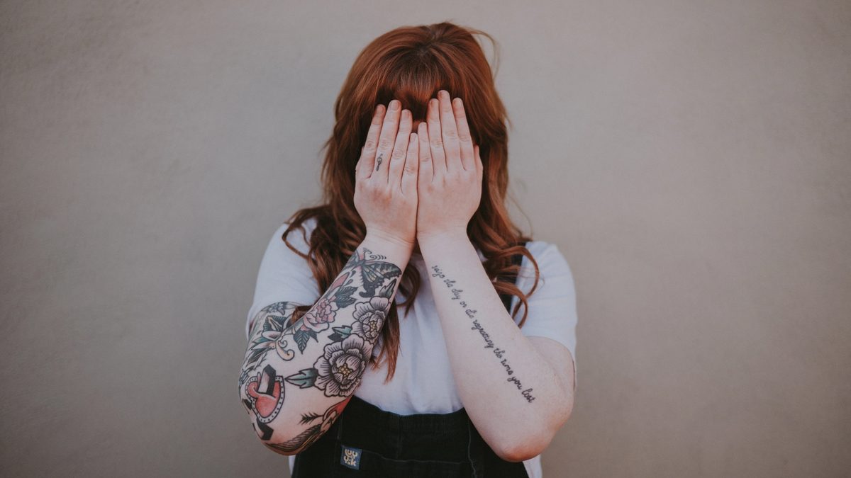 A person with long red hair and tattooed arms covers their face with their hands.