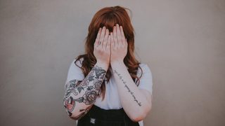 A person with long red hair and tattooed arms covers their face with their hands.