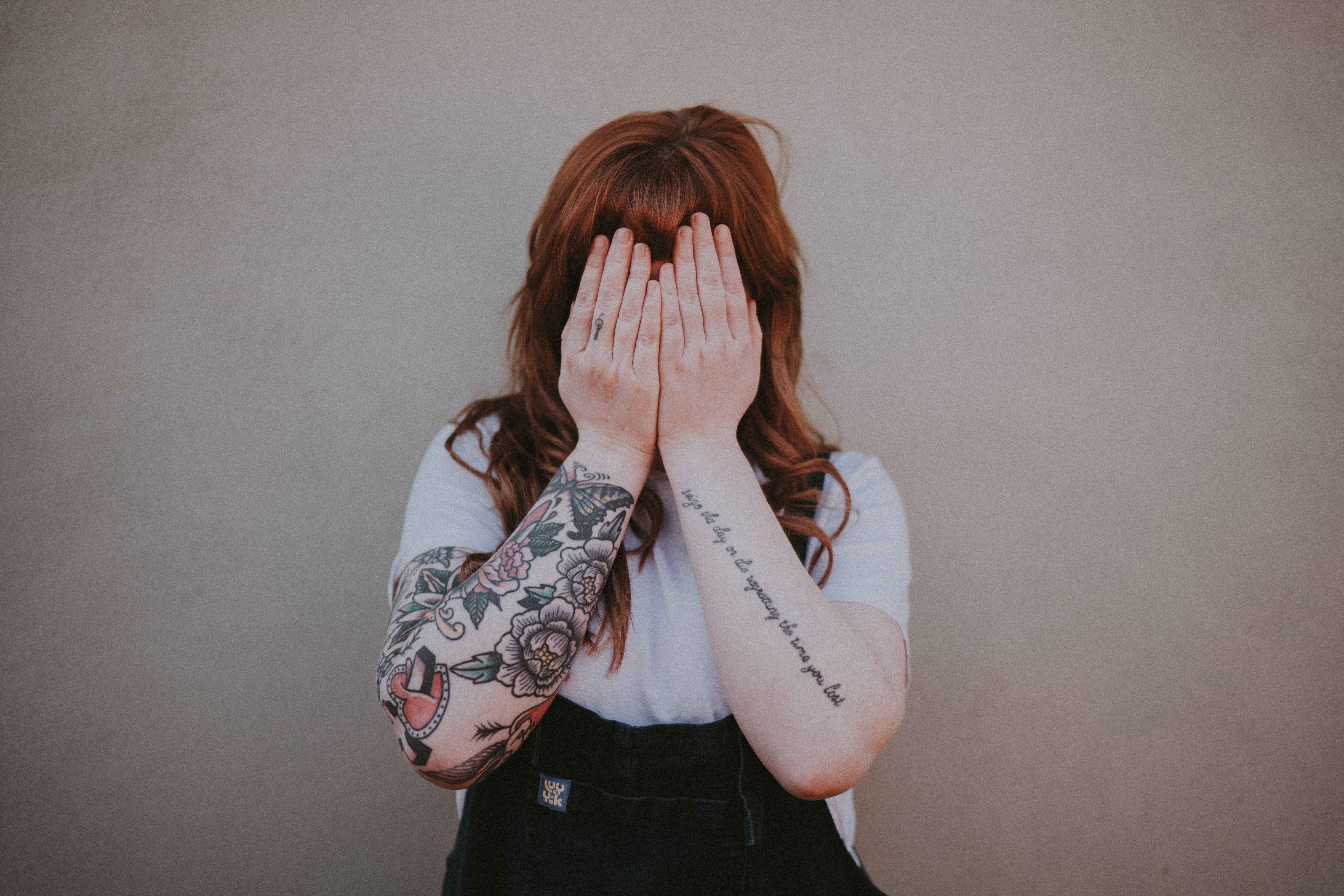 A person with long red hair and tattooed arms covers their face with their hands.