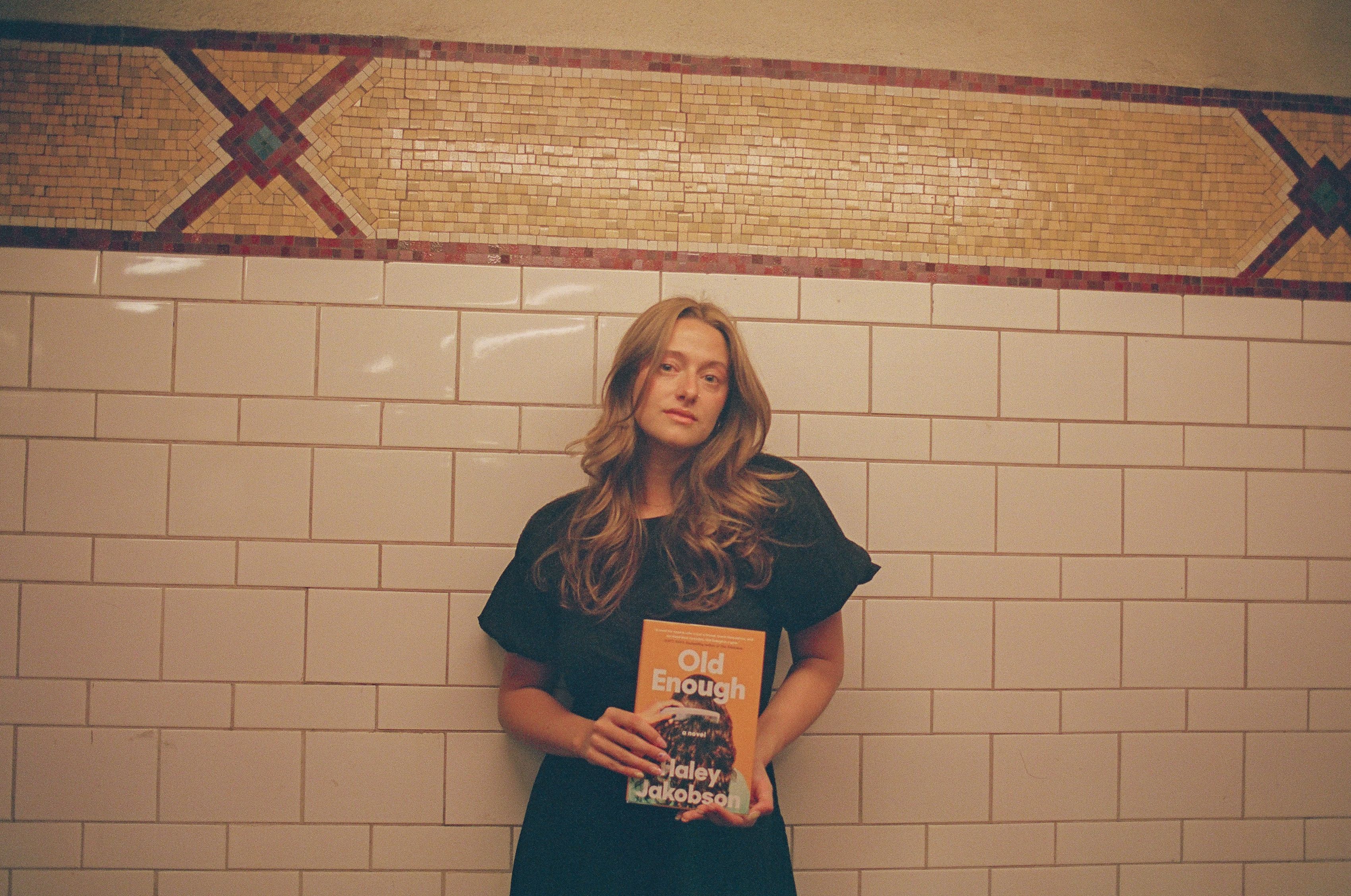 Haley Jakobson, a white queer femme, stands against an ornate tile backdrop while posing with her debut book, "Old Enough."